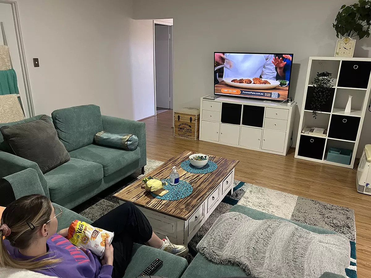 The second lounge room of Sudden Comfort's accommodation. A woman is playing sitting on the couch watching a cooking show while enjoying a snack. The room also has a bookshelf and house plants.
