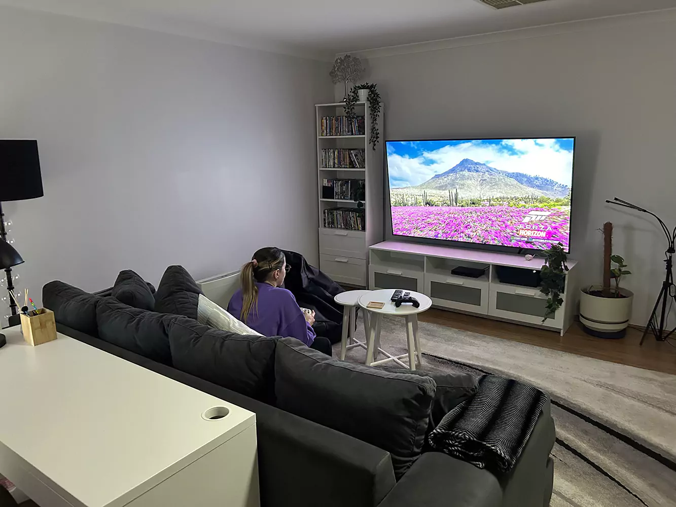 The main lounge room of Sudden Comfort's accommodation. A woman is playing a racing game on a playstation while sitting on the couch. The room also has a bookshelf and house plants.