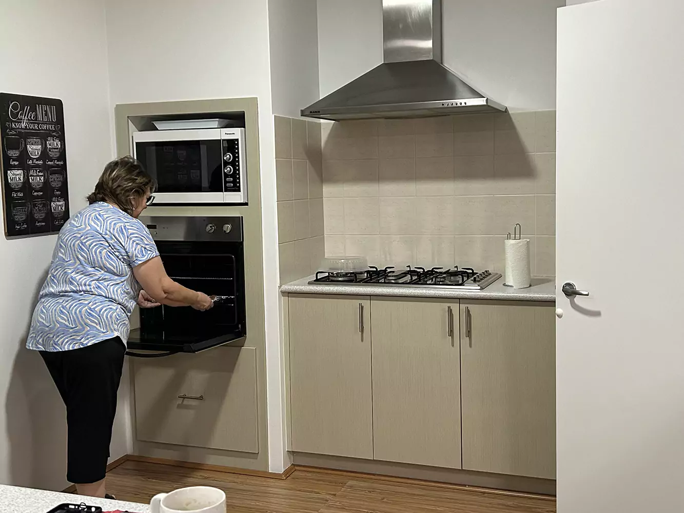 A woman is using the oven in the Sudden Comfort kitchen. She's removing a tray that may have freshly baked cookies. The kitchen is clean and spacious with modern appliances.