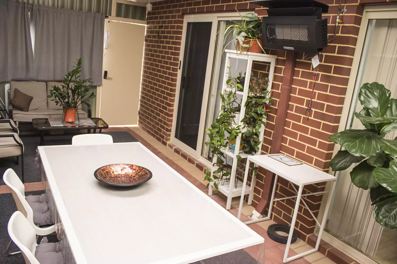 The image shows the outdoor patio of the Sudden Comfort's accommodation. There are many bright green house plants, heating options, seating a dining table and couch seating.