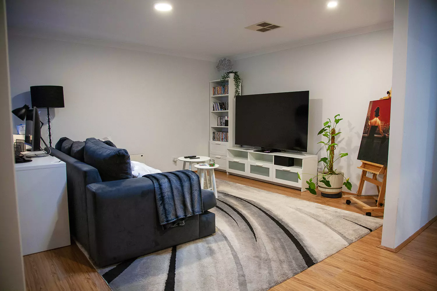 The image shows the lounge room area of the Sudden Comfort's accommodation. There is a book shelf, a large TV, house plants and a red painting of a woman in the corner. The room is inviting and well furnished with soft lighting and a comfortable lounge to sit on.
